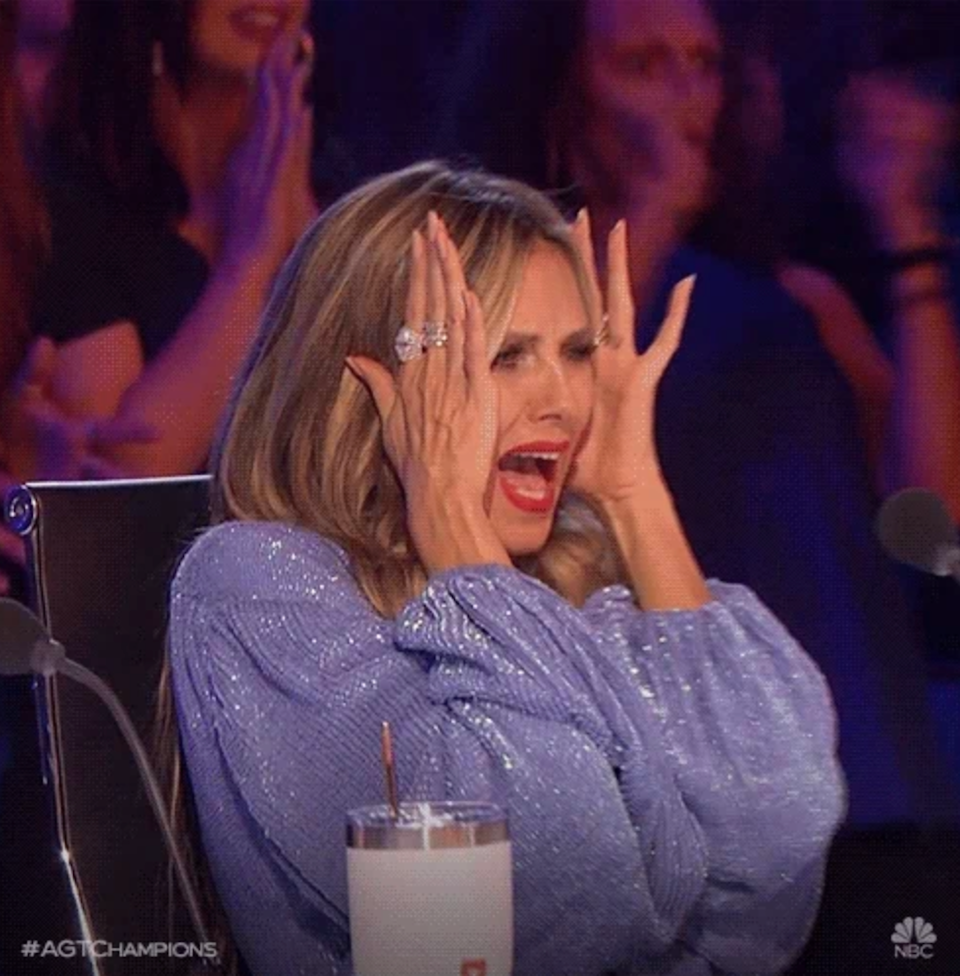 A person with long hair in a sparkly top expresses excitement or surprise, holding their face with both hands, sitting at a panel with a microphone