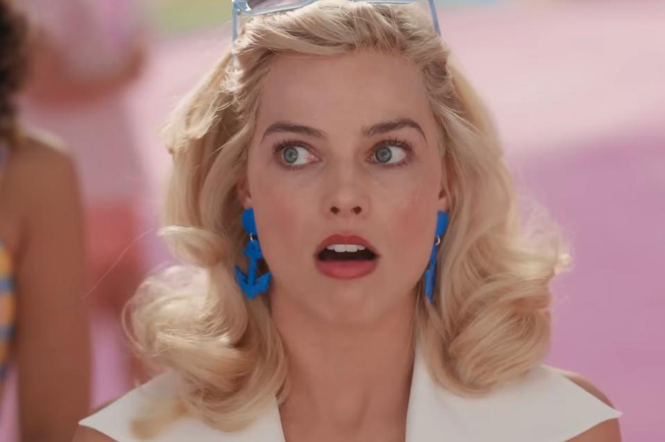 Person with wide-eyed expression, styled blonde hair, wearing blue geometric earrings, and a white top, appears surprised or in awe