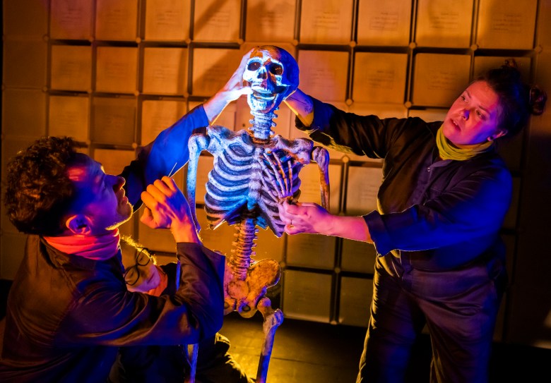 Two people in dark jumpsuits stand on either side of a life-size skeleton, which is lit from below. The face of another person is visible near ground level on the left.