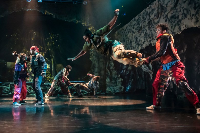 Three pairs of dancers onstage in dim light, engaged in what appears to be combat. A young woman and a man face off against each other on the left; he has a fist raised. Two men are center in the rear, one crouched in a defensive posture while the other holds what looks like a light sabre. On the right are two men, one in mid-flight while the other grabs at his ankles. They are all wearing grunge/punk type clothing.