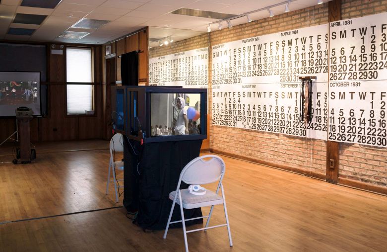 In the center of the gallery is a bloc known as the of two TV's screening videos back to back, with white folding chairs and headphones stationed in front of them. On the brick gallery wall to the right are large wall calendars from 1981 hung horizontally.