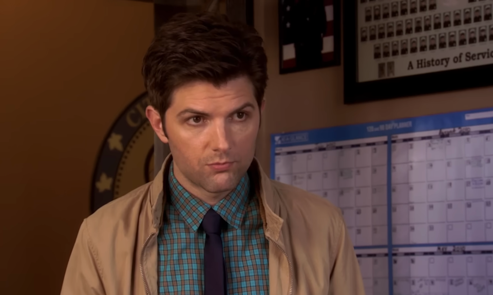A person with short hair wearing a checkered shirt and tie stands indoors, looking focused. A calendar is visible in the background