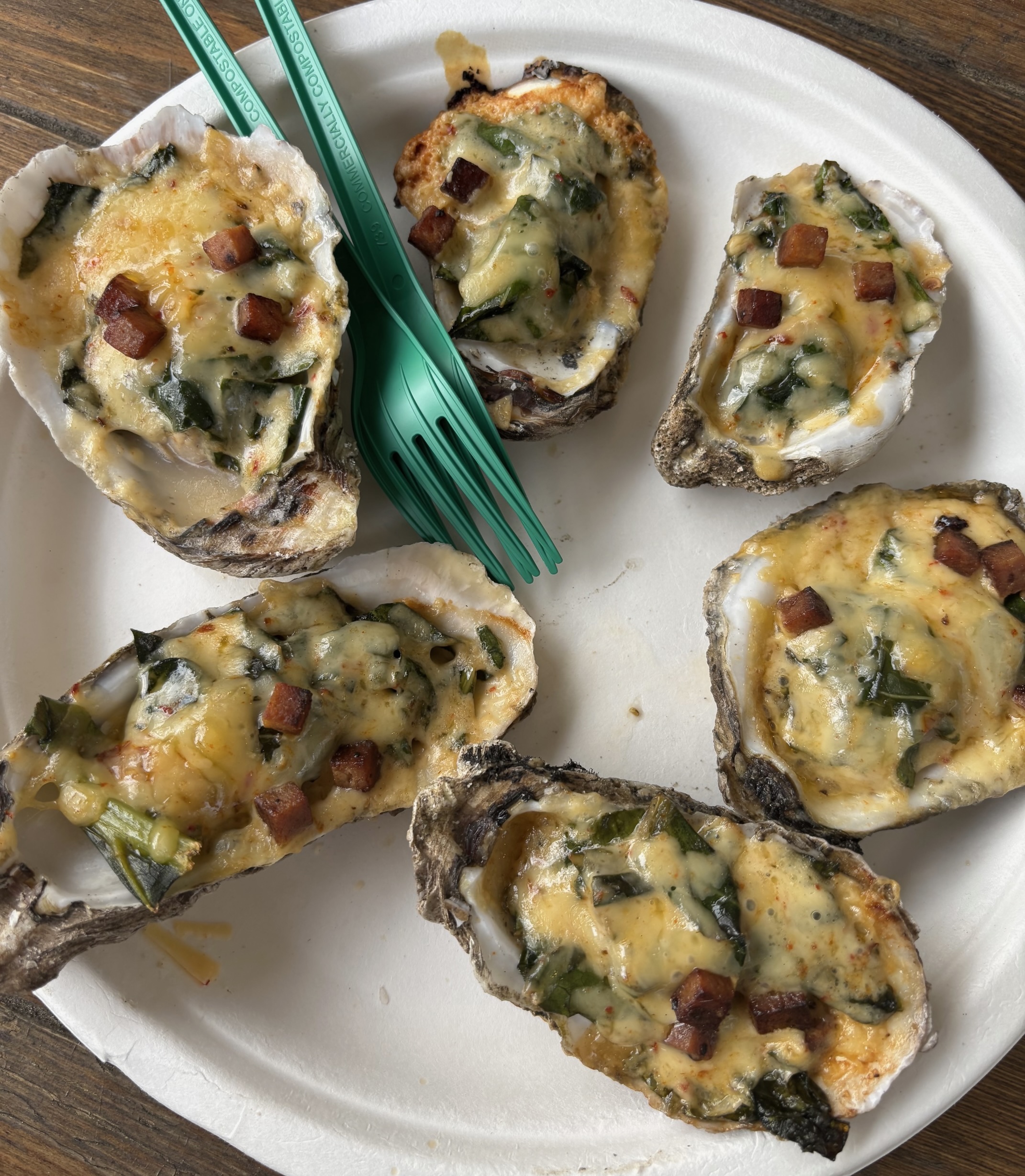 Six chargrilled oysters on a white paper plate topped with cheese, collards, and spam bits from Shuckboyz in Atlanta. Two green plastic forks are stacked on top of each other between two oysters. 